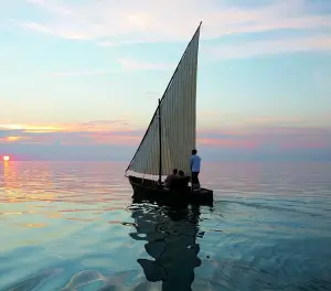 Traditional sailboat cruising across calm ocean waters during a colorful sunset in a tropical destination.