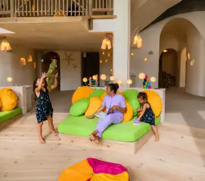 Children playing with a caregiver in a bright indoor family lounge featuring colorful beanbags, books, and a spacious play area.