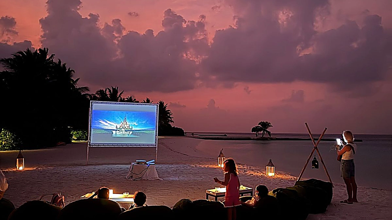 Outdoor beach cinema at sunset with cozy bean bags, lanterns, and guests watching a movie by the ocean