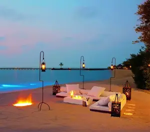 Private beachfront dining setup with lounge seating, lanterns, and a fire pit on a tropical beach at sunset.