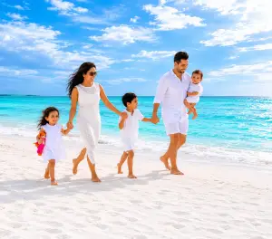 Family of five walking barefoot along a white sandy beach beside turquoise ocean waters under a dramatic blue sky.