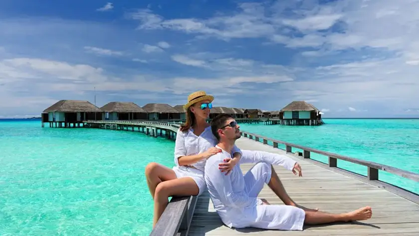 A couple in white resort wear sitting on a wooden jetty over vibrant turquoise water with tropical overwater bungalows in the distance.
