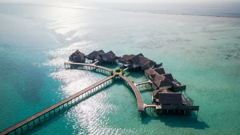 Aerial view of a secluded cluster of overwater villas connected by curved wooden walkways over clear turquoise tropical water.