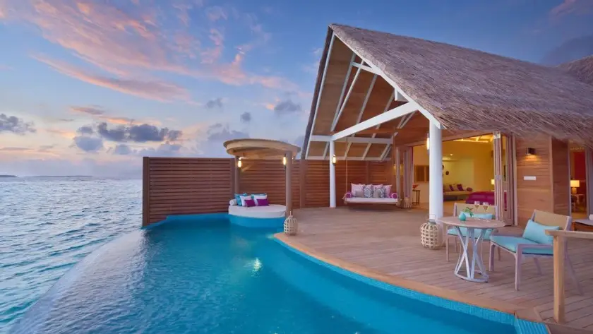 Overwater villa with a private infinity pool overlooking the ocean, surrounded by a spacious wooden deck and tropical-style furnishings beneath a colorful evening sky.