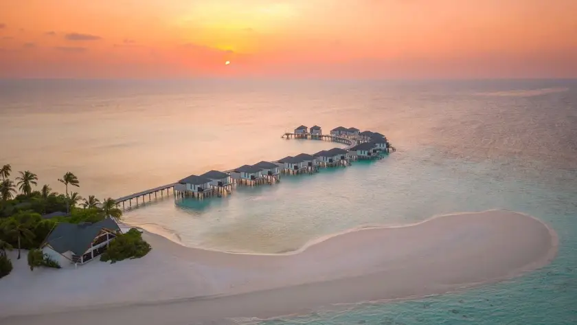 Aerial sunset view of a tropical island resort with overwater villas extending into calm turquoise sea beside a pristine white-sand beach.