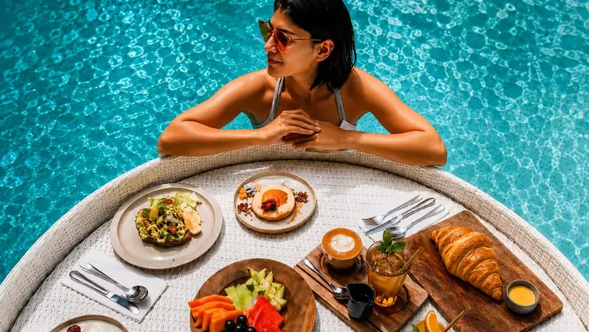 Woman relaxing in a swimming pool beside a floating breakfast tray with fruit, coffee and croissants