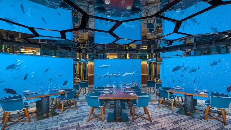 Underwater restaurant dining room with tables and blue chairs surrounded by large aquarium windows filled with tropical fish and sharks.