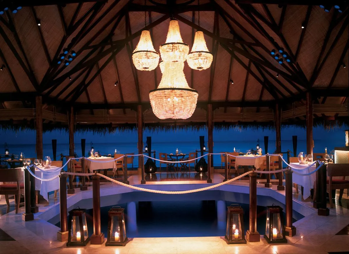 Elegant open-air oceanfront restaurant at dusk, featuring a large chandelier, candlelit tables, wooden beams, and views of the sea.