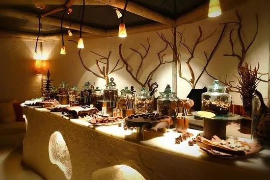 Warmly lit dessert buffet with cakes, chocolates, pastries, and jars of sweets displayed on a curved counter beneath decorative branch arrangements.