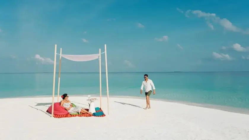Couple relaxing under a white canopy on a private Maldives sandbank with drinks and snacks, surrounded by clear turquoise water