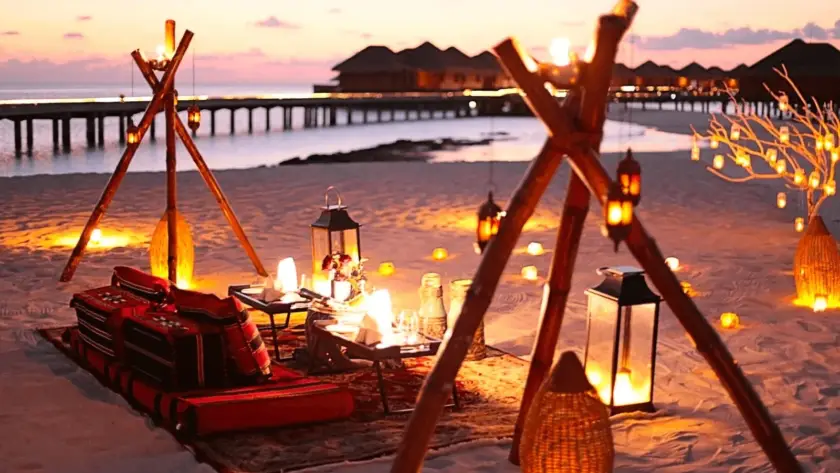 Romantic lantern-lit picnic setup on a Maldives beach at dusk, with red cushions, low tables and bamboo tripods, and overwater villas in the background