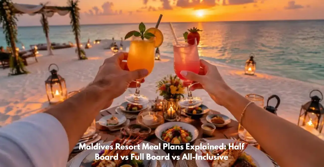 Two people toast colorful tropical cocktails over a beachfront dinner table at sunset in the Maldives, with lanterns, seafood, and turquoise ocean views.