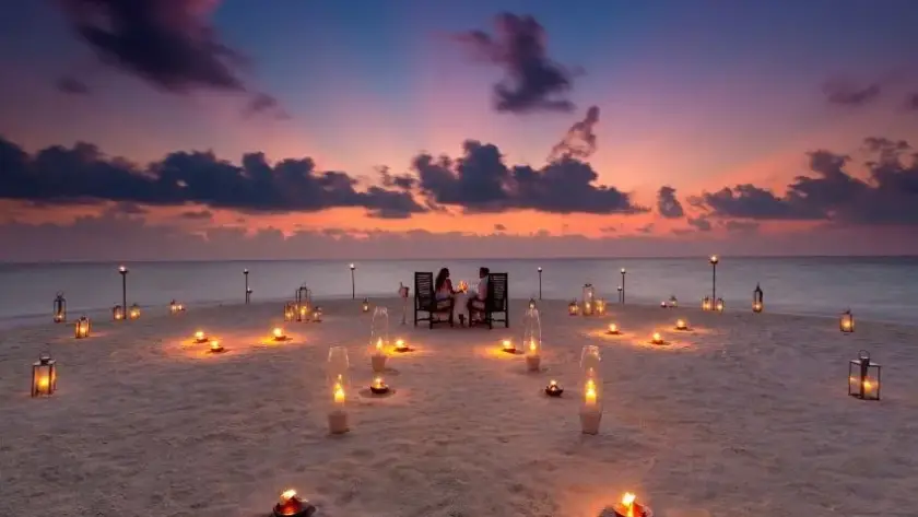 Couple enjoying a private candlelit sandbank dinner in the Maldives under a pink and purple sunset sky, ringed by lanterns